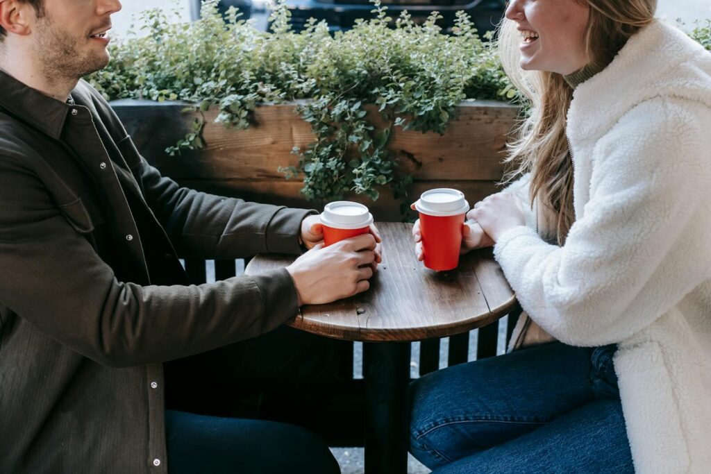 Crop faceless couple sitting at wooden table with paper cups of coffee and looking at each other while having date