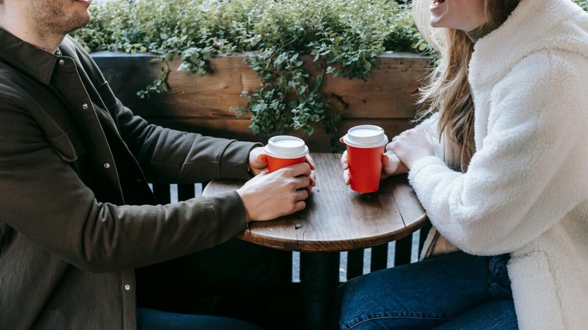 Crop faceless couple sitting at wooden table with paper cups of coffee and looking at each other while having date