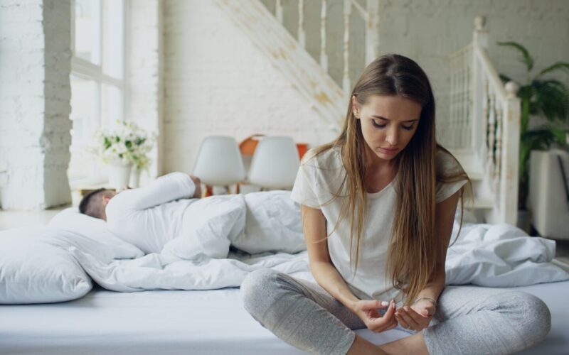 Woman sitting on bed, man sleeping behind her